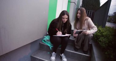 İki genç kız bir defter ve tablet kullanarak birlikte okuyorlar. Takım çalışmasını ve arkadaşlığı somutlaştırıp öğreniyorlar..