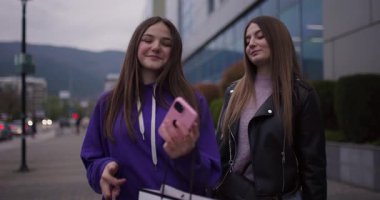 İki genç kız alışveriş yaptıktan sonra caddede yürür, telefonlarına bakar ve sohbet ederler. Satın almaktan memnun görünüyorlar..