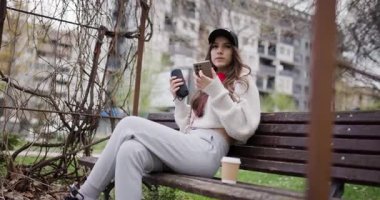 Genç Kadın Park Bench 'te Müzik ve Kahveden Hoşlanıyor: Smartphone ve Bluetooth Hoparlörüyle Sakin Bir Şehir Gevşeme Anı