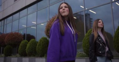 İki genç kız alışveriş çılgınlığının tadını çıkarıyorlar, çantalarını taşıyorlar ve modern bir binanın önünden geçerken sohbet ediyorlar..