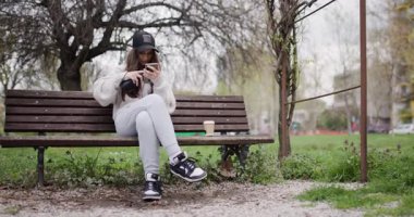 Akıllı Telefon Kullanan Genç Kız, Park Bench 'te kahve ve kulaklıkla dinleniyor.