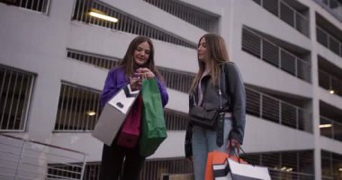 İki genç kız alışveriş merkezinin önünde durup aldıkları eşyalara bakıyor. Çantaları tutarlar, sohbet ederler ve birlikte gülerler..