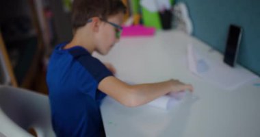 Gözlüklü genç bir çocuk odasında masasına oturur ve beyaz bir kağıt parçası katlar..