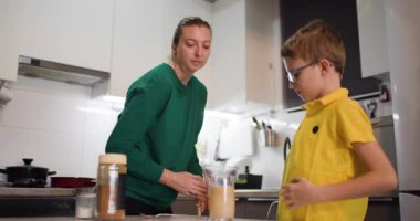 Genç bir çocuk dikkatle bir bardağa kahve ile süt dökerek lezzetli bir katmanlı içecek yaratıyor. Annesi gülümseyerek izliyor..