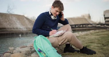 Parkta ders çalışan bir genç. Genç erkek öğrenci dışarıda öğreniyor, notlar alıyor ve telefonda konuşuyor..