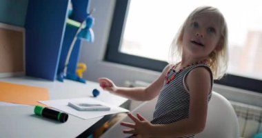 Şirin küçük bir kız masasında el sanatları ve el sanatları ile vakit geçirmekten hoşlanıyor. Bir kağıt parçasına boya basıyor ve boyalı ellerini gösteriyor..