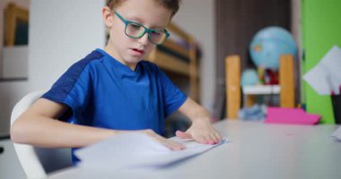 Gözlüklü genç bir çocuk masasında oturur ve dikkatle bir parça kağıt katlar. Harekete odaklanmış ve meşgul..