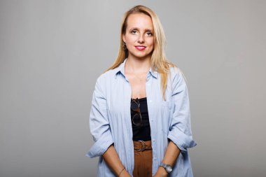 Bir iş kadını, siyah bluz ve kahverengi pantolon üzerine günlük mavi bir gömlek giyerek sıcak bir gülümseme ile güven içinde ayakta durur. Ortam minimalist, profesyonel ama rahat bir atmosfere katkı sağlıyor..