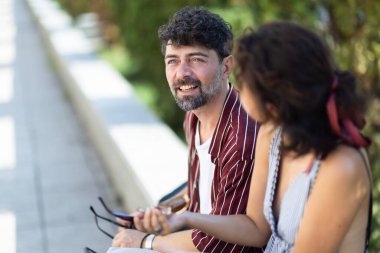 Güneşli bir günde, bir erkek ve bir kadın dışarıda dostça sohbet etmekten hoşlanırlar. Her ikisi de çizgili yaz kıyafetleri giyiyor ve rahatlamış, gülümseyen bir etkileşimi paylaşıyorlar..