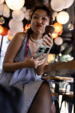 Bir kadın, renkli fenerlerle dolu rahat bir kafede dondurma külahı ve akıllı telefonuyla eğlenceli bir şekilde etkileşime girer ve canlı bir atmosfer yaratır..