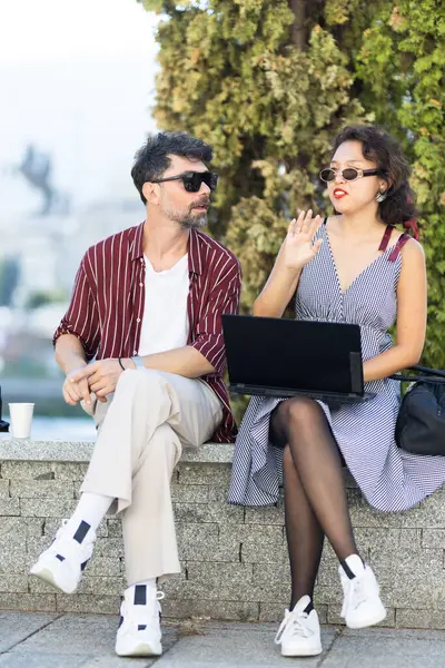 Güneş gözlüklü iki şık insan, yemyeşil bir ortamda güneşli bir günün tadını çıkarırken, şehir parkındaki bir bankta dizüstü bilgisayarla gündelik bir konuşma yapıyorlar..