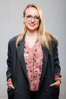 Profesyonel bir kadın şık gri bir ceketin altına şık pembe bir çiçek bluzu giyiyor. Çağdaş bir ofis ortamında güven içinde gülümsüyor, başarıyı ve gücü somutlaştırıyor..