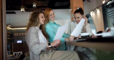 Üç iş kadını canlı bir kafede toplanarak yeni projeleri tartışırken, belgeleri gözden geçirip görüşlerini paylaşıyorlar. Takım çalışmaları, profesyonel bir ortamda destek ve yaratıcılık gösteriyor..