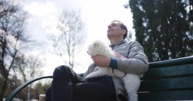 Bir adam küçük beyaz bir köpeği kollarında sallarken parkta bir bankta dinleniyor. Güneş, rüzgarda hafifçe sallanan ağaçlar gibi parlıyor. Huzurlu bir atmosfer yaratıyor..