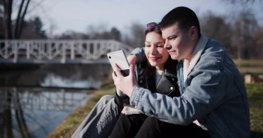 Güneşli bir günde, genç bir çift nehir kenarında bir parkta selfie çekiyor. Gülümsüyorlar ve birbirlerine eşlik etmekten zevk alıyorlar..