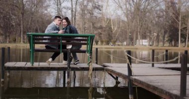 Genç bir çift gölün üzerindeki bankta selfie çekiyor. Birlikte eğlenirken gülümser ve öpüşürler..