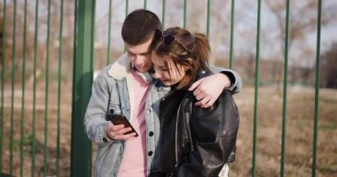 Genç bir çift çitin yanında durup birlikte telefona bakıyor. Sevgiyle sarılırlar, güneşli bir günde birbirlerine eşlik ederek eğlenirler..