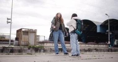 Sırt çantalı ve kameralı iki genç kadın, yaklaşan maceraları için heyecanlı bir şekilde tren istasyonunda seyahat planlarını tartışıyor..