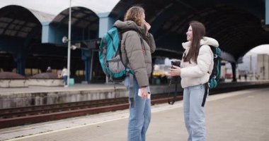 İki genç bayan arkadaş tren istasyonunda bekliyor, kabarık ceketler ve sırt çantaları giyiyorlar, gülümsüyorlar ve kameradaki seyahat fotoğraflarına bakıyorlar..