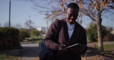Genç bir Afrikalı Amerikalı adam tabletine bakarken parkta yürüyor. Gülümsüyor ve ekrandaki bir şeye tepki veriyor..