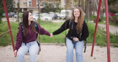 İki genç kadın salıncakta oturup el ele tutuşup gülüyorlar. Biri kamerayla fotoğraf çekiyor. Parkta güneşli bir günde arkadaşlıklarının tadını çıkarıyorlar..