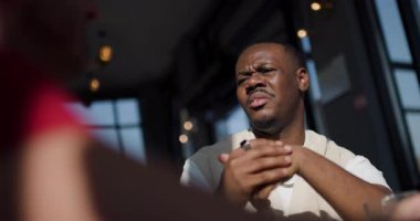 Bir adam bir masada oturur, karşısındaki birini dikkatle dinler, güneşli bir kafede..