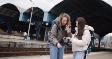 Sırt çantalı iki genç kadın bir tren istasyonundalar. Biri diğerinin fotoğrafını çekiyor. Gülüyorlar ve yolculukları için heyecanlı görünüyorlar..