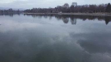 Low aerial drone footage of Treska lake in Skopje, North Macedonia during an autumn day with leafless trees and cloudy skies.