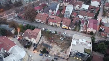 Aerial view of a construction site in an urban area with excavators and machinery, surrounded by buildings, streets, and parked cars.