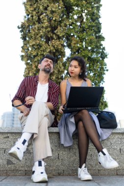Two stylish individuals sit on a stone bench, collaborating with a laptop outdoors. The setting is casual and bright, promoting teamwork.