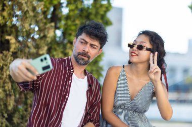 A couple enjoys a sunny day, taking a selfie together. The woman is making a peace sign, and the man is focused on the camera, with green trees behind them.