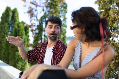 Two people having a lively conversation outdoors, surrounded by greenery on a sunny day, capturing a moment of friendship and communication.