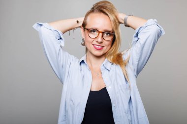 A confident businesswoman stands with her hands behind her head, wearing a light blue shirt and glasses. Her casual yet professional style reflects her vibrant personality and ready attitude.