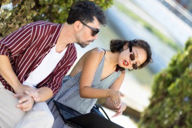A man and woman having a casual conversation outdoors. Both are wearing sunglasses on a sunny day, seated by the water.