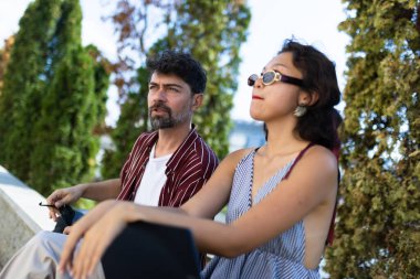 Two friends sitting outdoors in a park, engaging in conversation on a sunny day. The scene captures a relaxed and serene atmosphere with trees in the background.