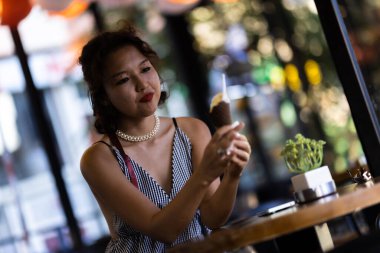 A woman in a cafe enjoys an ice cream cone, creating a relaxed and joyful atmosphere. She wears a striped dress and a pearl necklace, surrounded by soft lighting and a cozy setting.