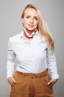 A confident businesswoman stands with her hands in her pockets, dressed in a white shirt and brown pants, radiating professionalism and self-assurance in a bright space.