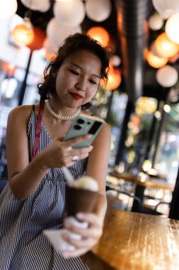 A young woman with red lipstick and pearl necklace relaxes with ice cream in a trendy cafe, using her smartphone.