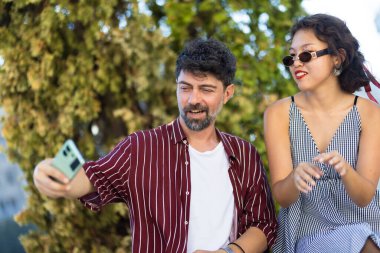 A young couple enjoying a sunny day outdoors, taking a selfie with a smartphone, and capturing joyful, fun moments together.