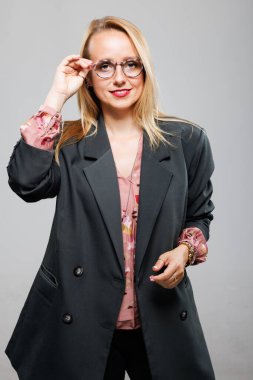 A businesswoman dressed in a stylish dark blazer and floral blouse stands confidently while adjusting her glasses. She smiles warmly, embodying professionalism and elegance in a studio environment.