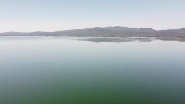 Serene lake panorama with reflections of the sky and mountains. Calm water surface and distant village. Peaceful natural landscape.