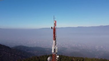 Telecommunication tower with antennas and satellite dish against a backdrop of a misty cityscape and snow-capped mountains.