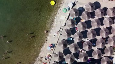 Beach umbrellas and lounge chairs by Lake Ohrid create a tranquil oasis where swimmers, sunbathers and tourists enjoy the beautiful day. The people play in the lake or relax on the pebbled beach.