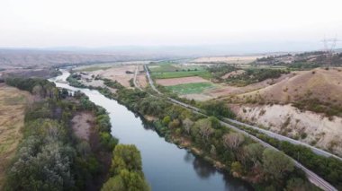 Aerial view of the Vardar River in North Macedonia, alongside a road and railway line, surrounded by lush green vegetation and rolling hills.