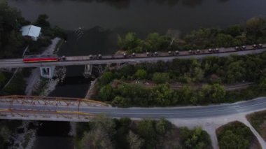 Aerial view of a red train crossing a bridge over a river, alongside a road bridge at sunset.