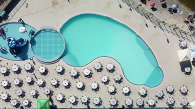An aerial view of a kidney-shaped swimming pool, round kiddie pool, and rows of round tables arranged for an event beside a lake.