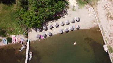 Rotating aerial footage from Lake Dojran in North Macedonia. People relax and swim by beach with straw umbrellas, boats and docks near road.