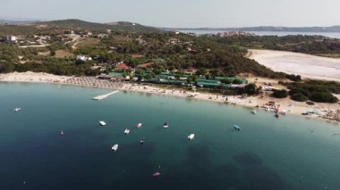 Ammouliani Island beach camp with clear turquoise waters, beach umbrellas, boats, a pier, and green trees in Greece.