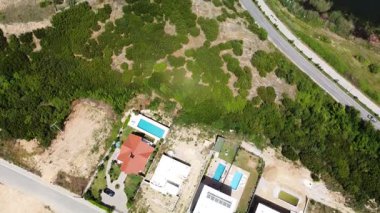 Aerial view of modern villas with swimming pools near a scenic road and a lake, showcasing a luxurious lifestyle amidst natural beauty.