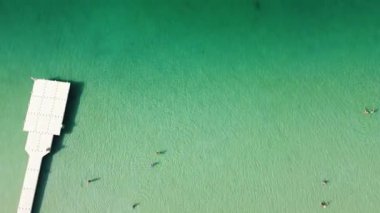 An aerial view of the clear turquoise waters of Ammouliani, Greece. People swim near a pier with boats anchored further out.